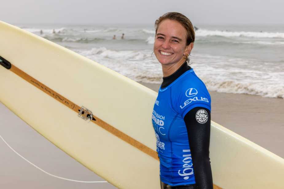 A smiling surfer in a blue wetsuit stands on the beach holding a surfboard, with ocean waves and other surfers in the background.