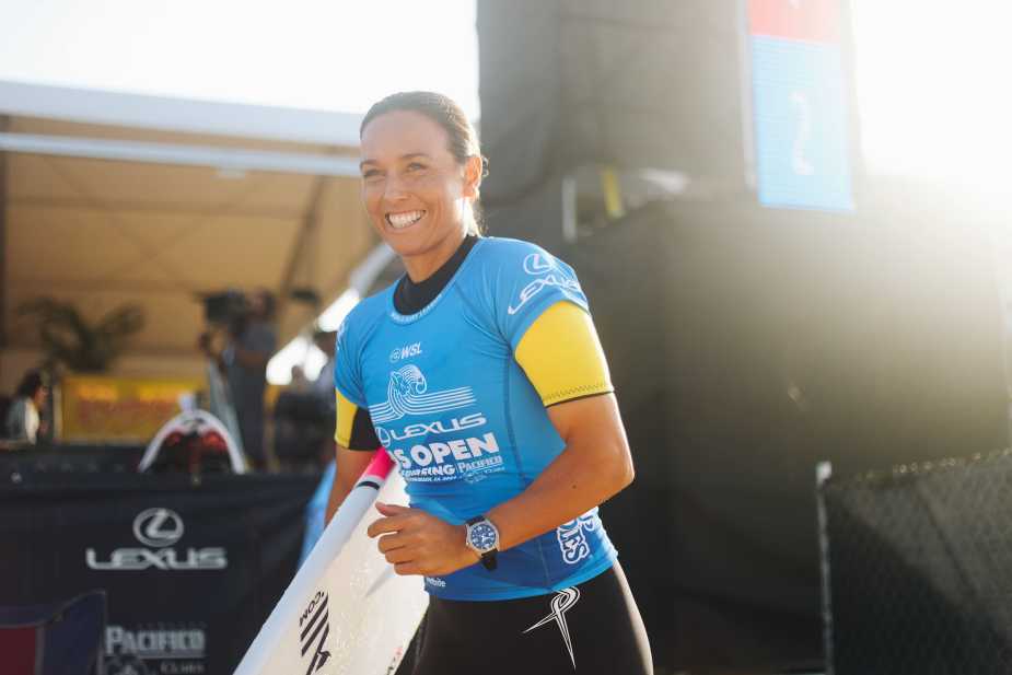 A smiling female surfer in a blue wetsuit stands holding her surfboard, with a bright backdrop of the beach and competition area.