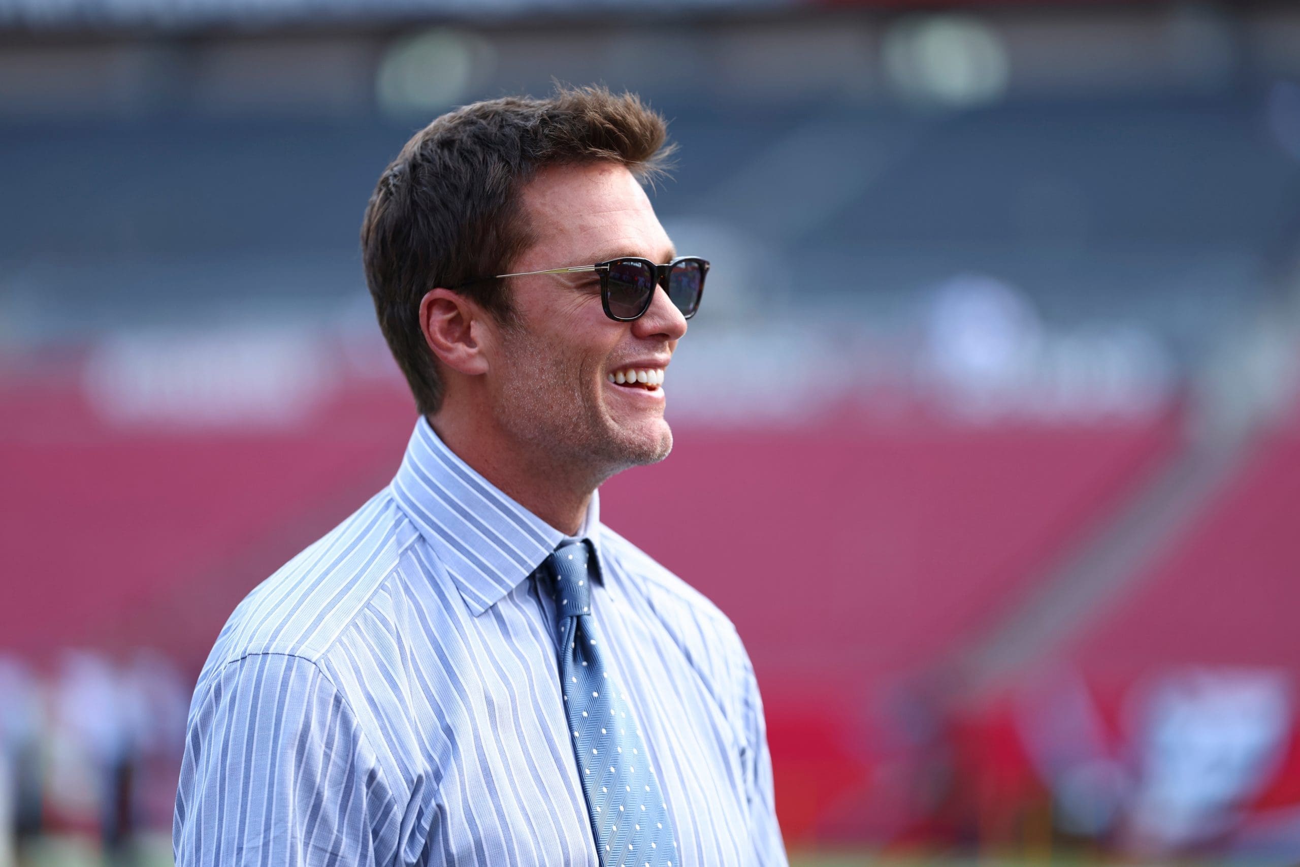 Tom Brady smiling in blue striped dress shirt and sunglasses at NFL stadium