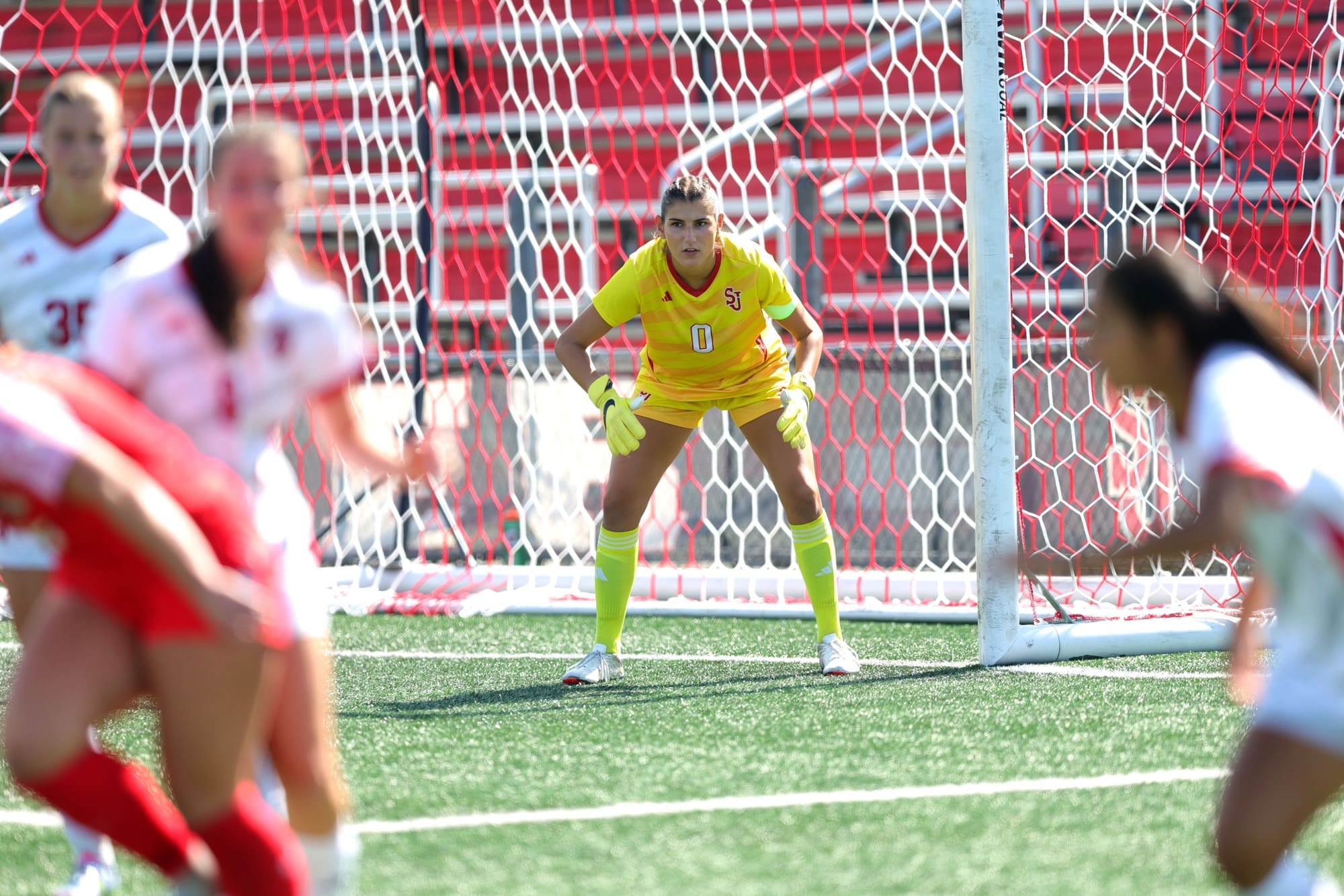 St. John's University women's soccer goalkeeper Kayla Bower in yellow uniform positioned in goal during a match, focused and ready to make a save with players in motion in the foreground
