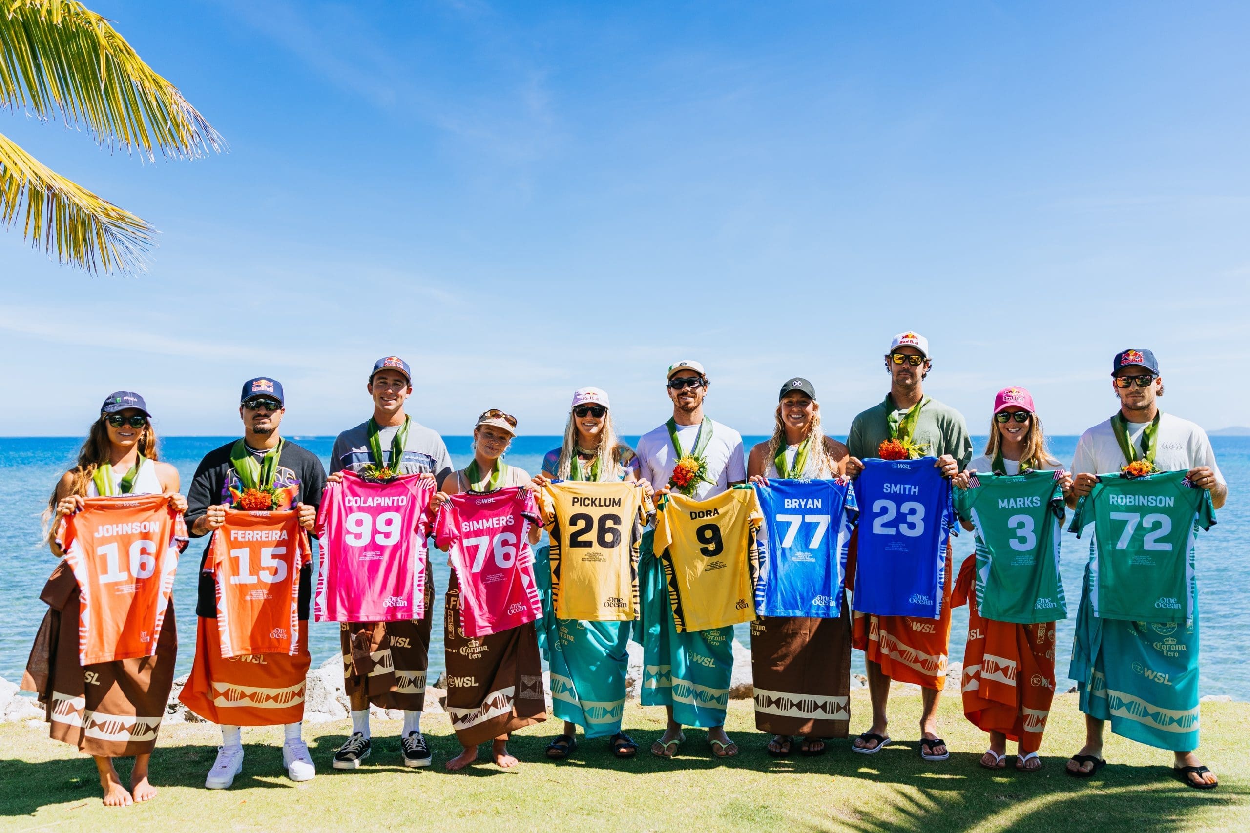 Ten surfers, including Bettylou Sakura Johnson, Italo Ferreira, Griffin Colapinto, Caitlin Simmers, Molly Picklum, Yago Dora, Gabriela Bryan, Jordy Smith, Caroline Marks, and Jack Robinson, stand side by side in front of the ocean at Cloudbreak, Tavarua, Fiji. Each holds a jersey with their name and number during a press conference before the 2025 Lexus WSL Finals.