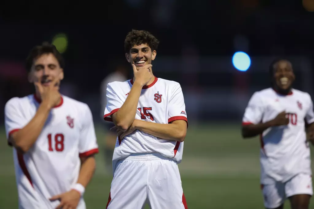 Three St. John's University soccer players celebrate history together on the field after scoring a goal, showing team unity and excitement during their match