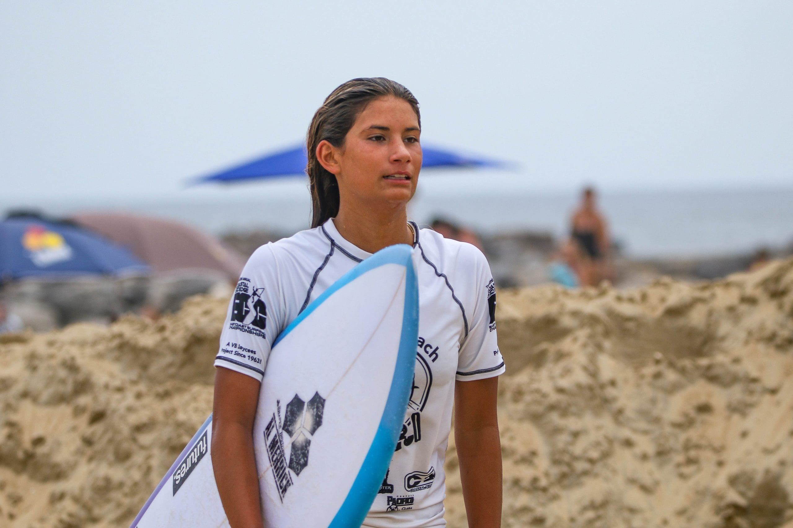 13-year-old surfer Teagan Meza holds white and blue surfboard while posing on beach during 2025 Virginia Beach Pro competition