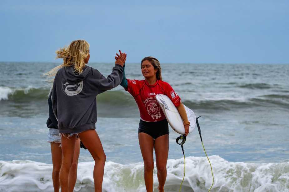 Zoey Kaina in red competition jersey receives a high-five from a supporter on Virginia Beach after her semifinal heat at the 2025 Virginia Beach Pro