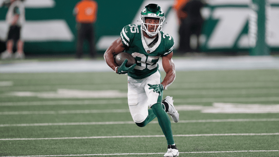 A running back in a green football uniform running on an NFL field while holding a football.