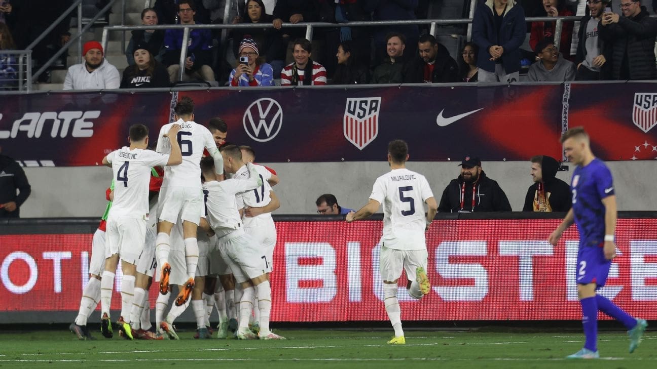 Serbia national team players in white jerseys celebrate goal in group huddle while USMNT player in blue walks away during international friendly at BMO Stadium
