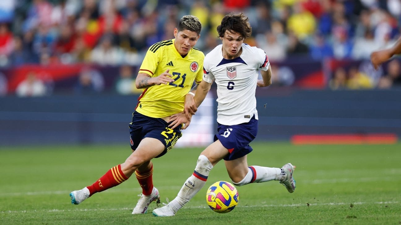 Paxten Aaronson number 6 in white USMNT jersey dribbles ball while being challenged by Colombia player in yellow jersey during international friendly