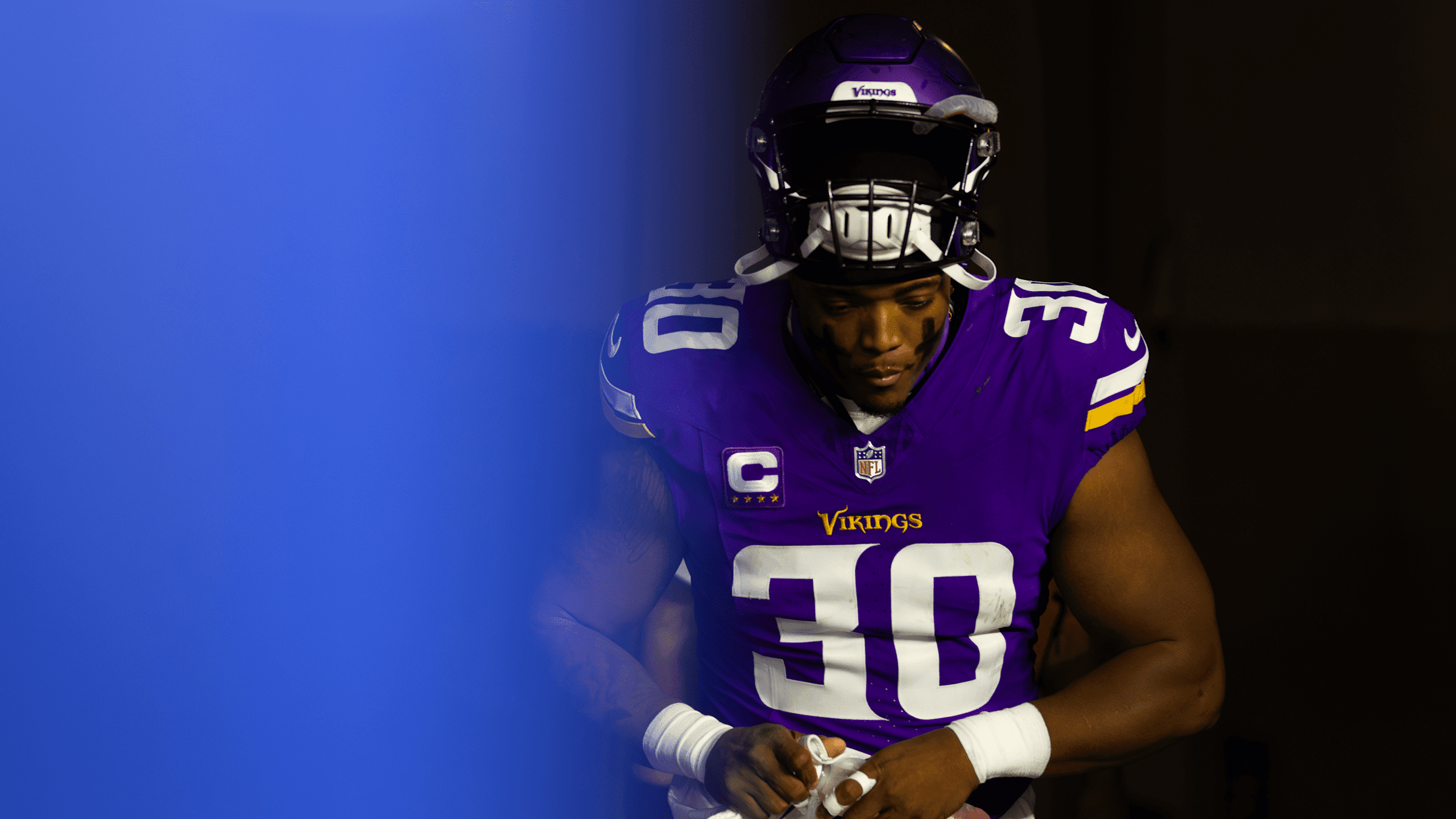 C.J. Ham in Minnesota Vikings purple home uniform with captain's "C" patch, holding a football in dramatic lighting with blue background