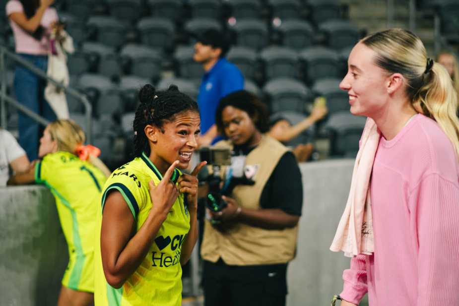 Croix Bethune, in a yellow CVS Health jersey, animatedly talking with Esme Morgan, in a pink top, after the Washington Spirit match, stadium seating in the background