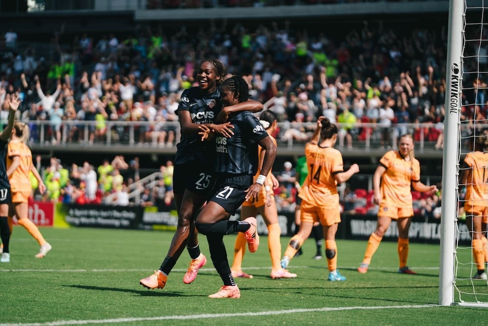 Washington Spirit players in black jerseys celebrate near the goal, with Gift Monday embracing a teammate after scoring, surrounded by Houston Dash players in orange uniforms.