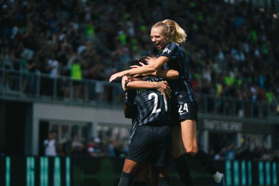 Washington Spirit players in black jerseys embrace in celebration, with a player wearing jersey number 21 being hugged by a teammate with blonde hair, packed stadium in background