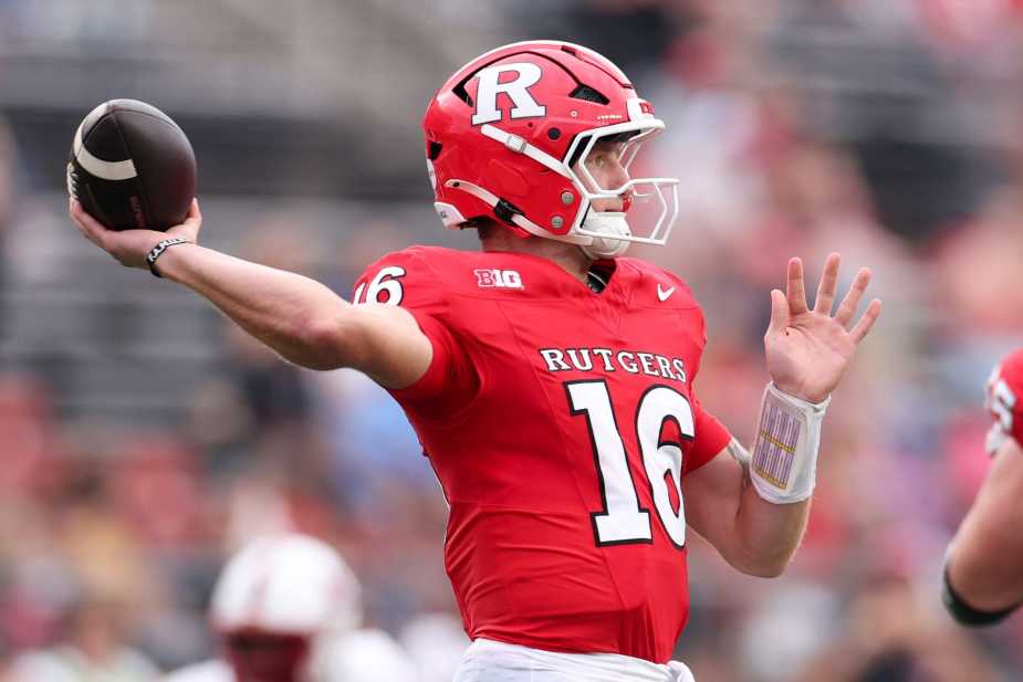 Rutgers quarterback Athan Kaliakmanis in red jersey number 16, holding a football and signaling during a game, wearing a bright red helmet with the Rutgers 'R' logo