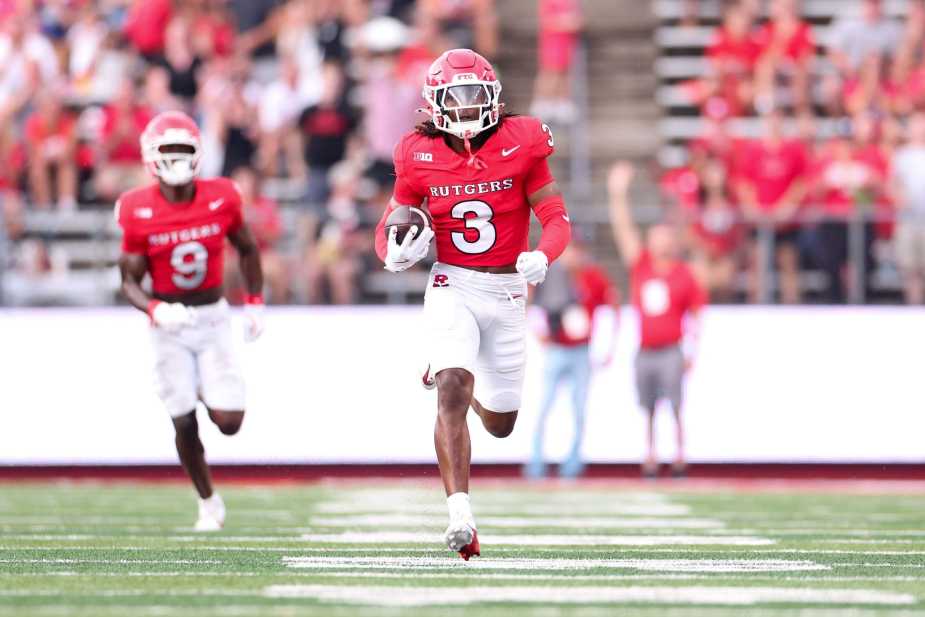 Rutgers Defensive back Bo Mascoe (#3) sprints down the field in a red Rutgers jersey, with teammate in background, against a blurred stadium crowd