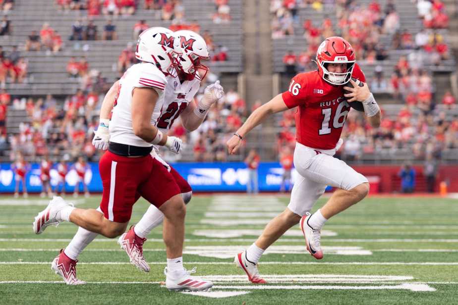 Rutgers quarterback Athan Kaliakmanis (#16) in red jersey scrambles to the right while being pursued by two Miami (Ohio) defenders in white jerseys