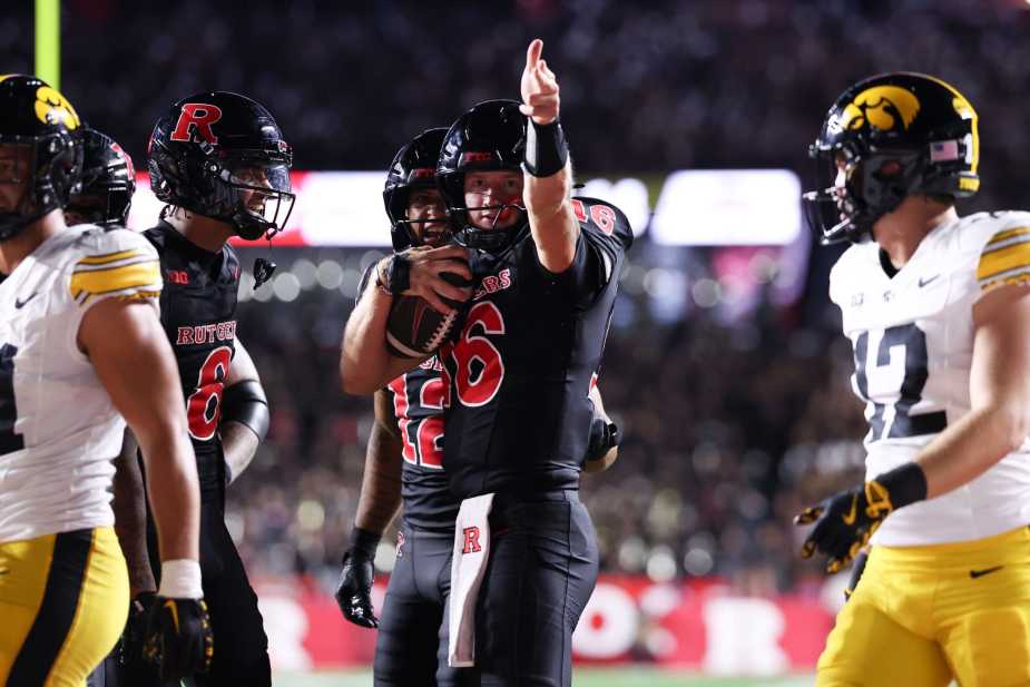 Rutgers quarterback Athan Kaliakmanis, number 16, signals first down while holding the football, surrounded by Iowa defenders in their white and gold uniforms during the night game