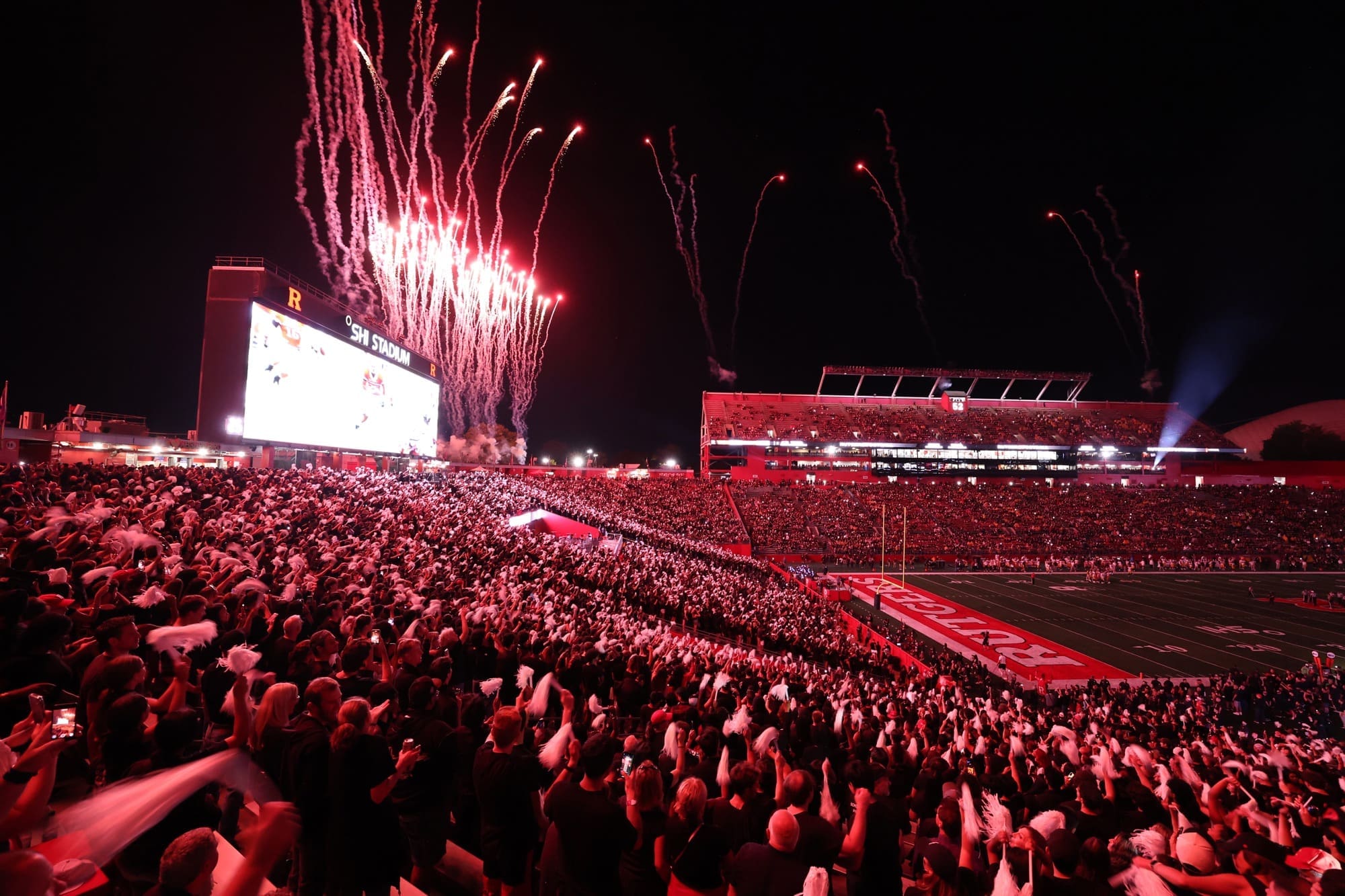 Spectacular fireworks display erupts over SHI Stadium during Rutgers football game, with packed stands of fans in red lighting and the iconic "R" logo visible on the stadium structure