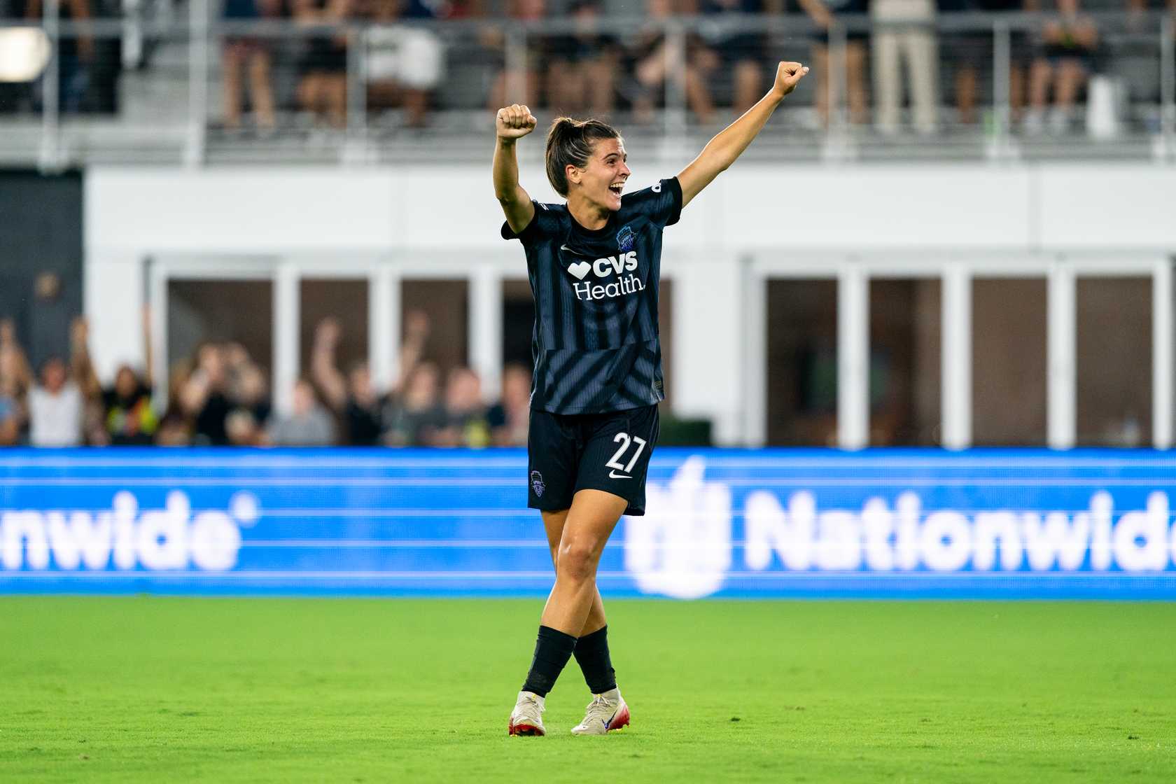 Sofia Cantore #27 of Washington Spirit celebrates with raised fists after a goal, wearing a black CVS Health jersey, standing on soccer field with Nationwide advertising board in background