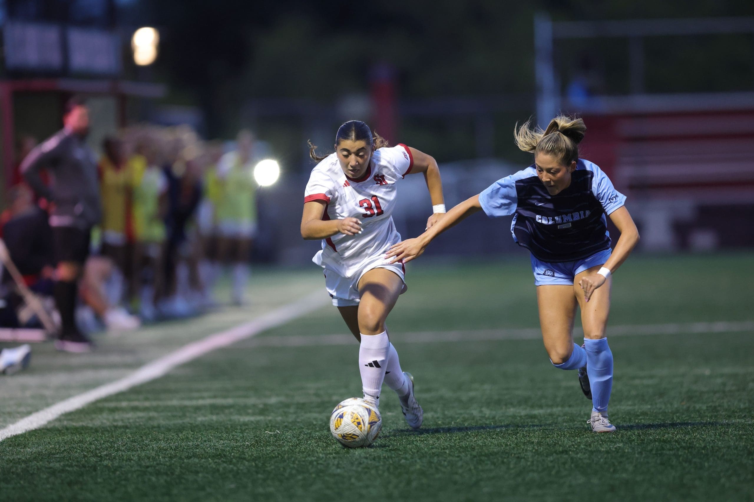 St. John's University forward Madison Mariani in white jersey with number 31 dribbles down the sideline while being pursued by a Columbia defender in light blue during women's college soccer match