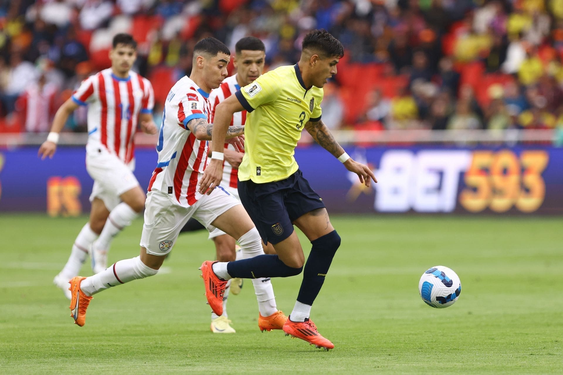 Ecuador player in yellow jersey dribbles past Paraguay defender in red and white striped kit during CONMEBOL World Cup qualifying match at Estadio Defensores del Chaco