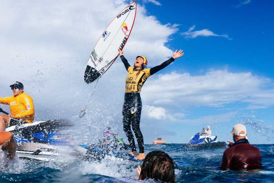 Molly Picklum celebrating her 2025 WSL World Championship victory in the ocean, holding her Rip Curl surfboard high above her head with arms outstretched, wearing her yellow championship jersey, surrounded by team members and water spray
