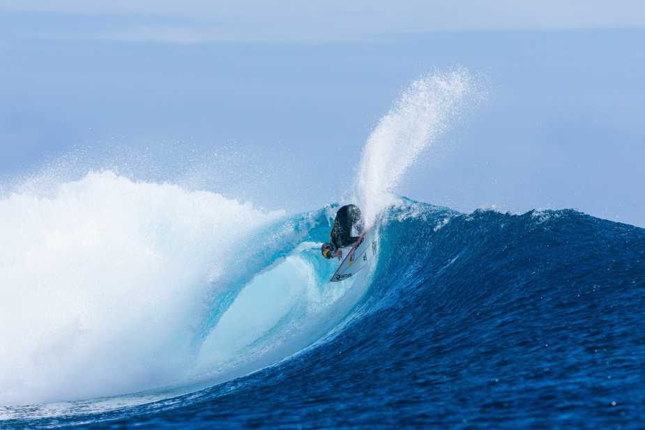 Molly Picklum executing a powerful surfing maneuver on a massive blue wave at Cloudbreak, Fiji, creating a dramatic spray of water