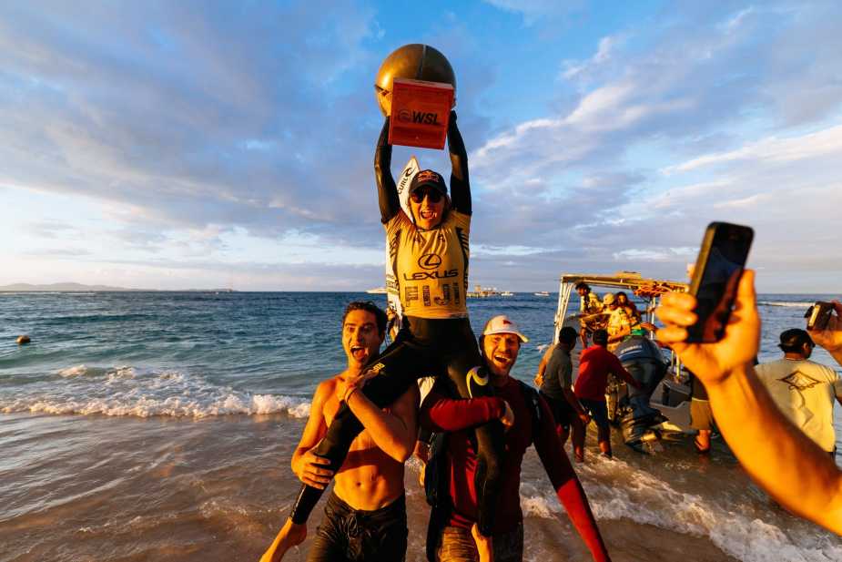Molly Picklum celebrating her World Surf League Championship, standing atop supporters on the beach, holding her WSL trophy high with excitement