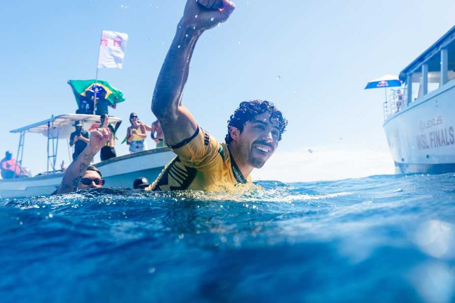 Yago Dora celebrating his 2025 WSL World Championship in the ocean, raising his fist with a Brazilian flag in the background, surrounded by support boats