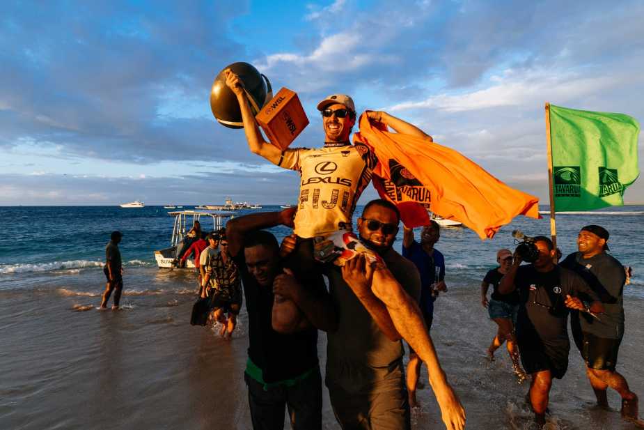Yago Dora being carried on supporters' shoulders on the beach, holding his WSL Championship trophy high above his head while wearing his yellow Lexus Fiji jersey, with Brazilian flag and Tavarua Island flag visible in celebration
