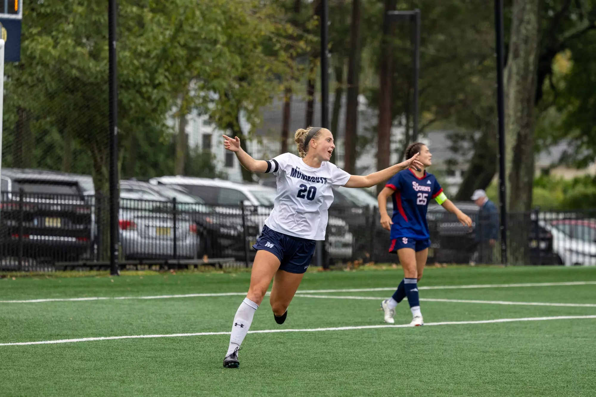 Monmouth Hawks forward #20 Summer Reimet celebrates with arms outstretched after scoring a goal, wearing white home uniform while a St. John's player in blue looks on in the background