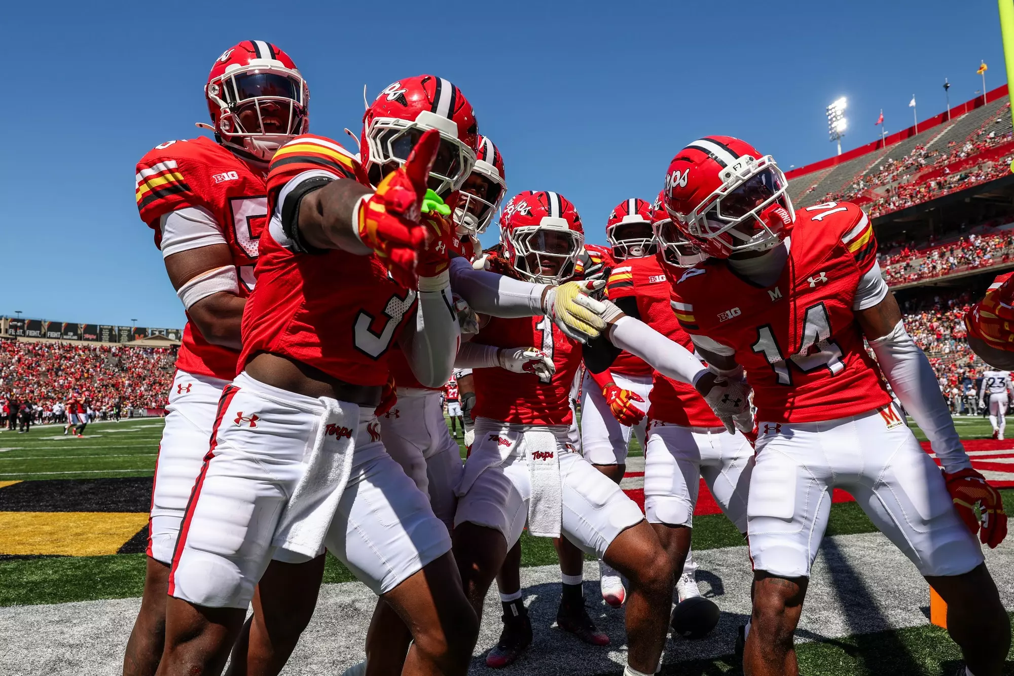 Maryland football players in red and white uniforms celebrate together in the end zone after scoring a touchdown, with teammates gathering around and pointing in celebration at SECU Stadium