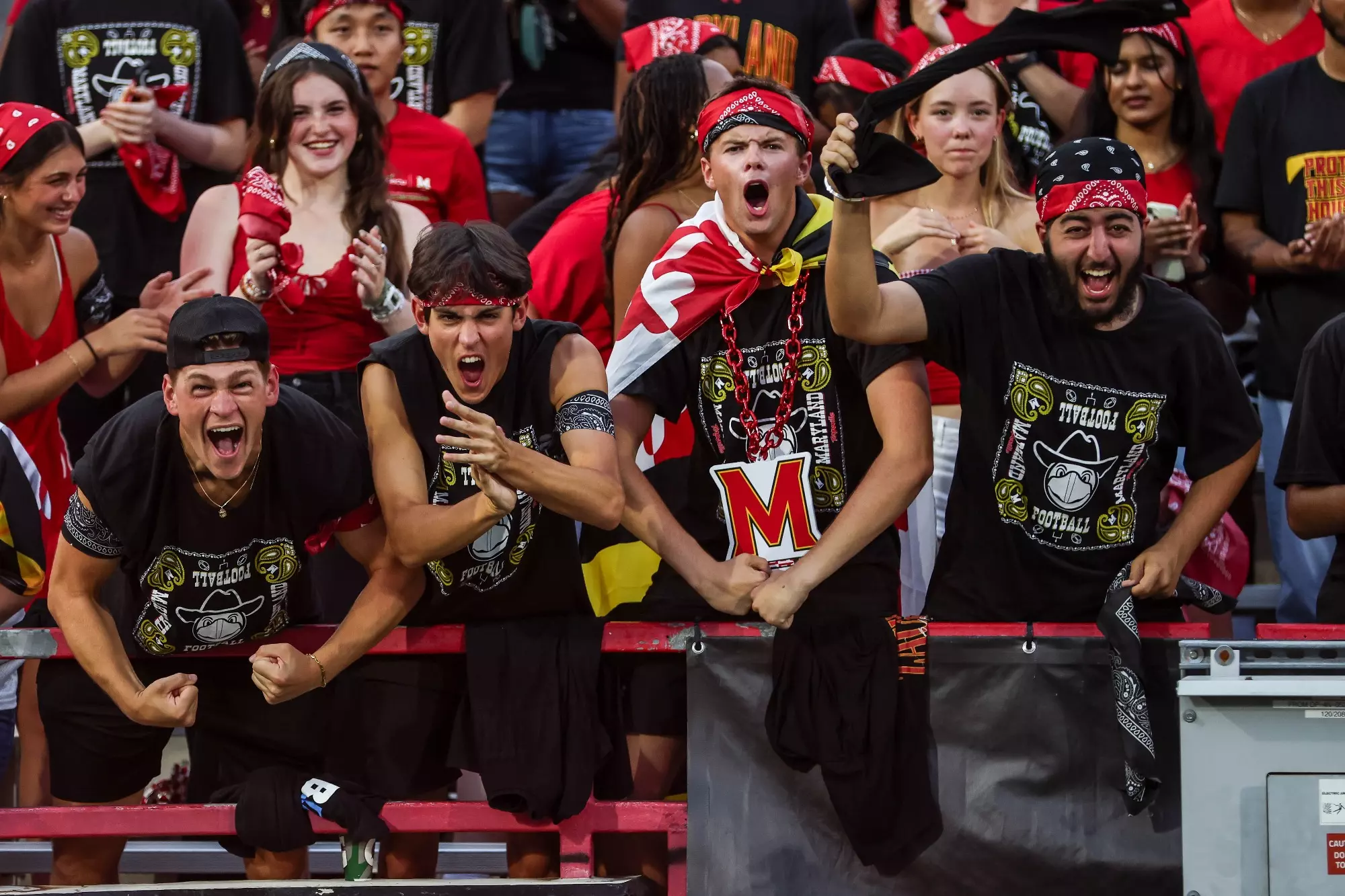 Enthusiastic Maryland football fans in the stadium stands cheer wildly, wearing red bandanas and black shirts with team logos, showing intense excitement and school spirit