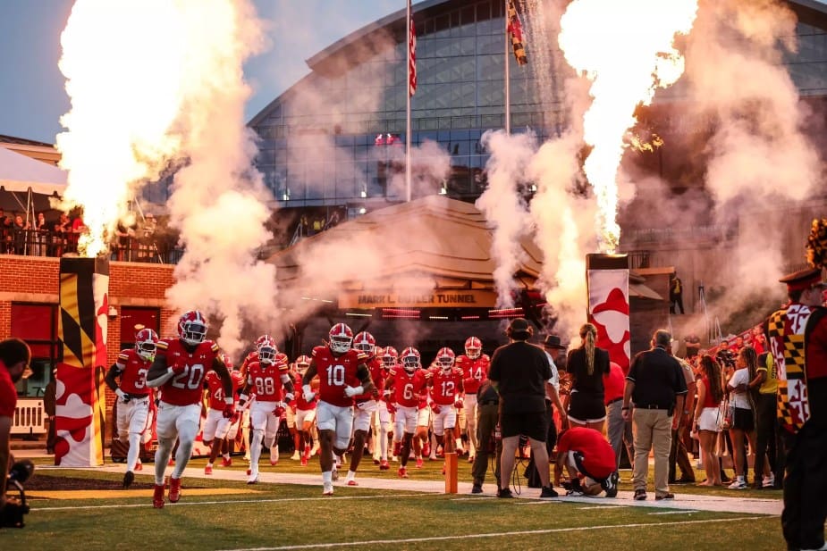 Maryland football team charges out of the Mark L. Butler Tunnel onto the field amid dramatic fire and smoke effects, with players in red uniforms running toward the camera