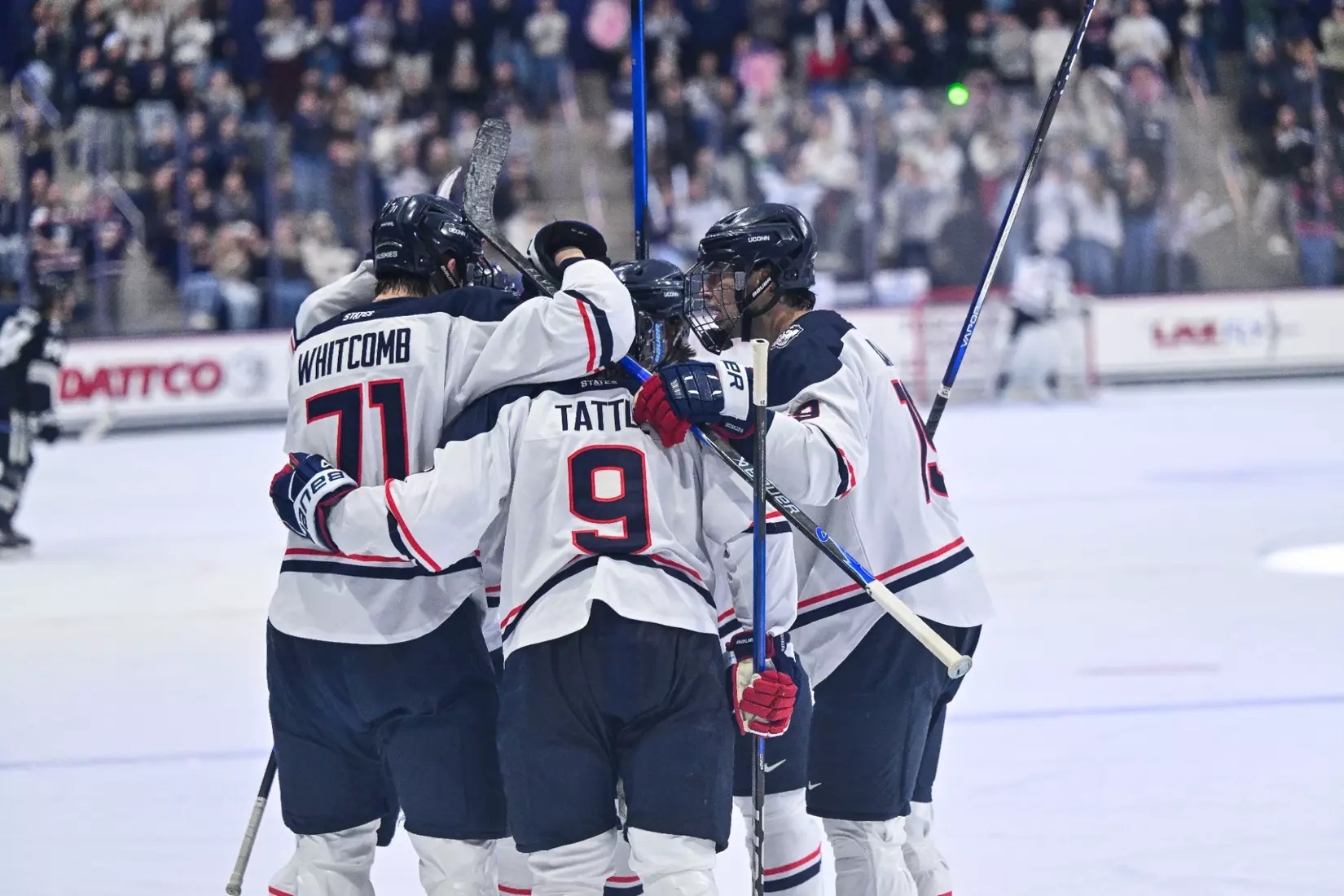 UConn men’s hockey players celebrate a goal on home ice, gathering in a group hug with visible player names and numbers, as the crowd cheers in the background.