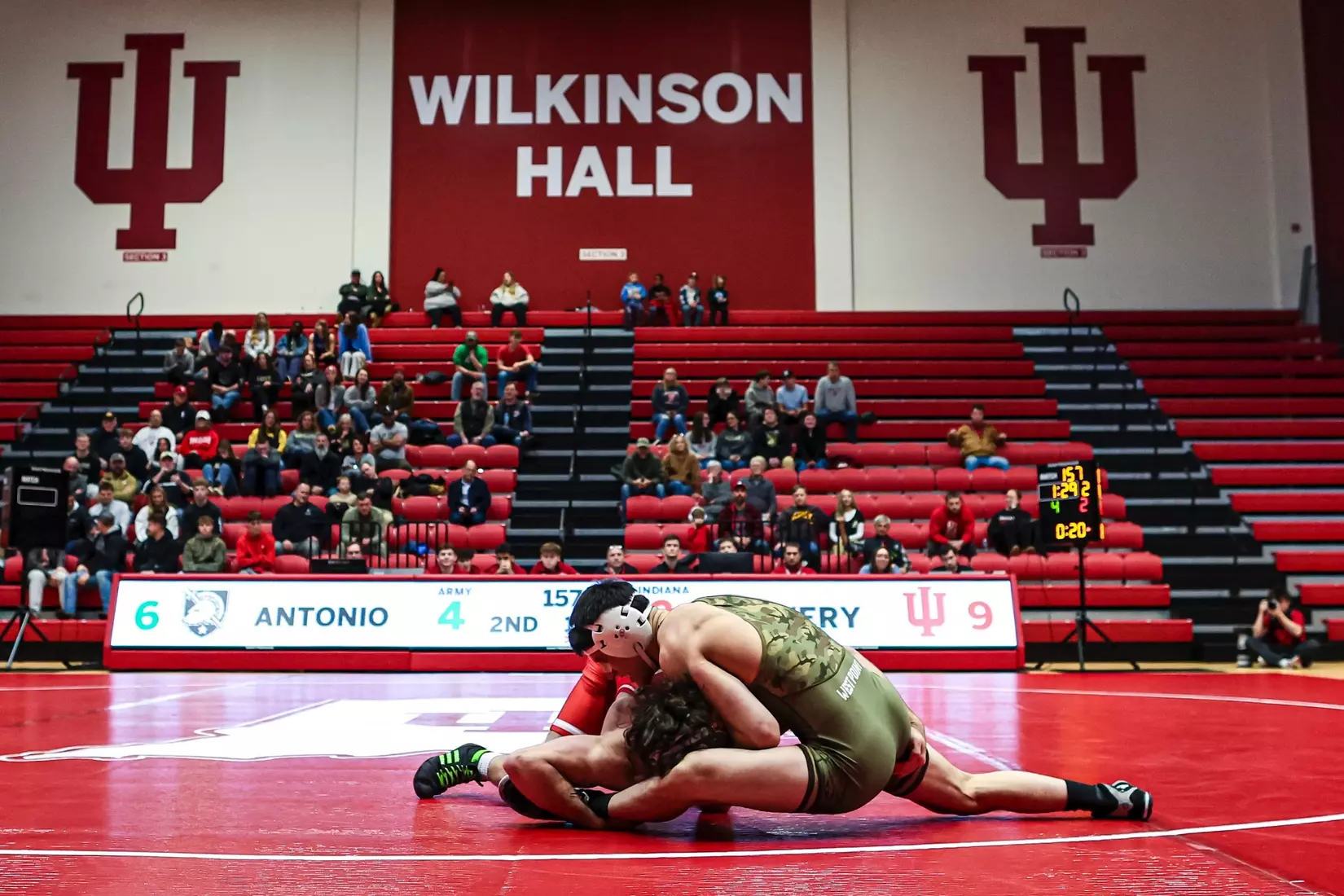 Army West Point wrestler in camouflage singlet grapples with an Indiana opponent on the mat at Wilkinson Hall, with spectators watching from red bleachers in the background.