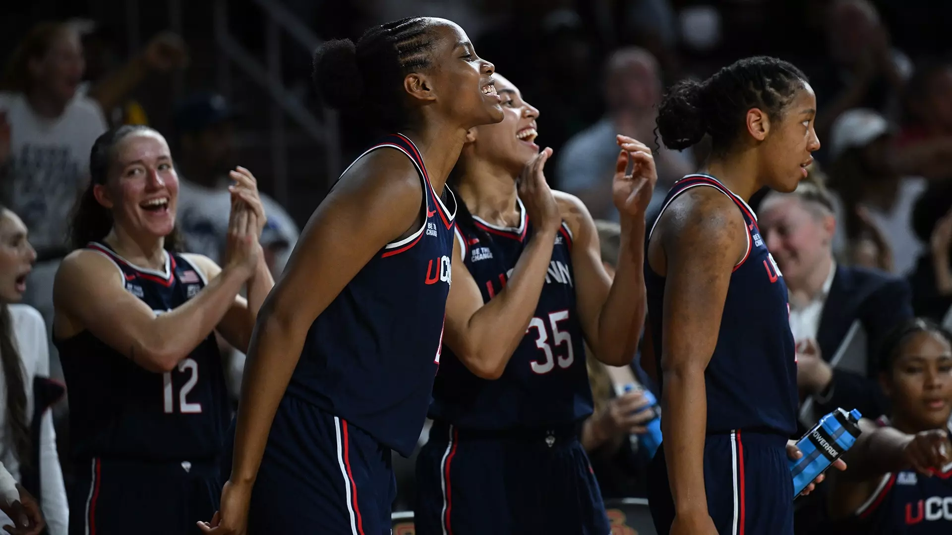 UConn women’s basketball players in navy uniforms celebrate and cheer from the bench during a game, smiling and clapping with excitement. Mandatory Credit: UConn Athletics