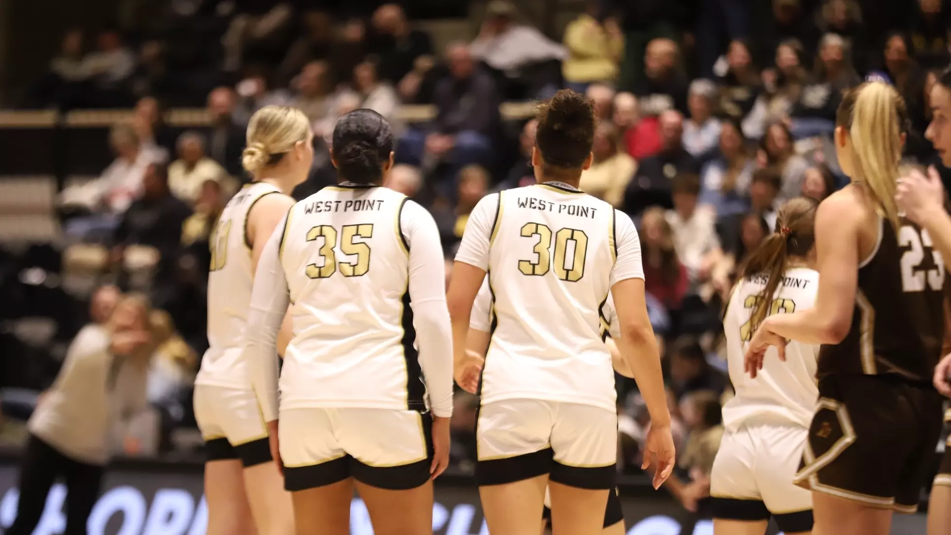 Army West Point women’s basketball players, wearing white uniforms with “WEST POINT” and jersey numbers visible, stand on the court during a home game as fans watch from the stands.