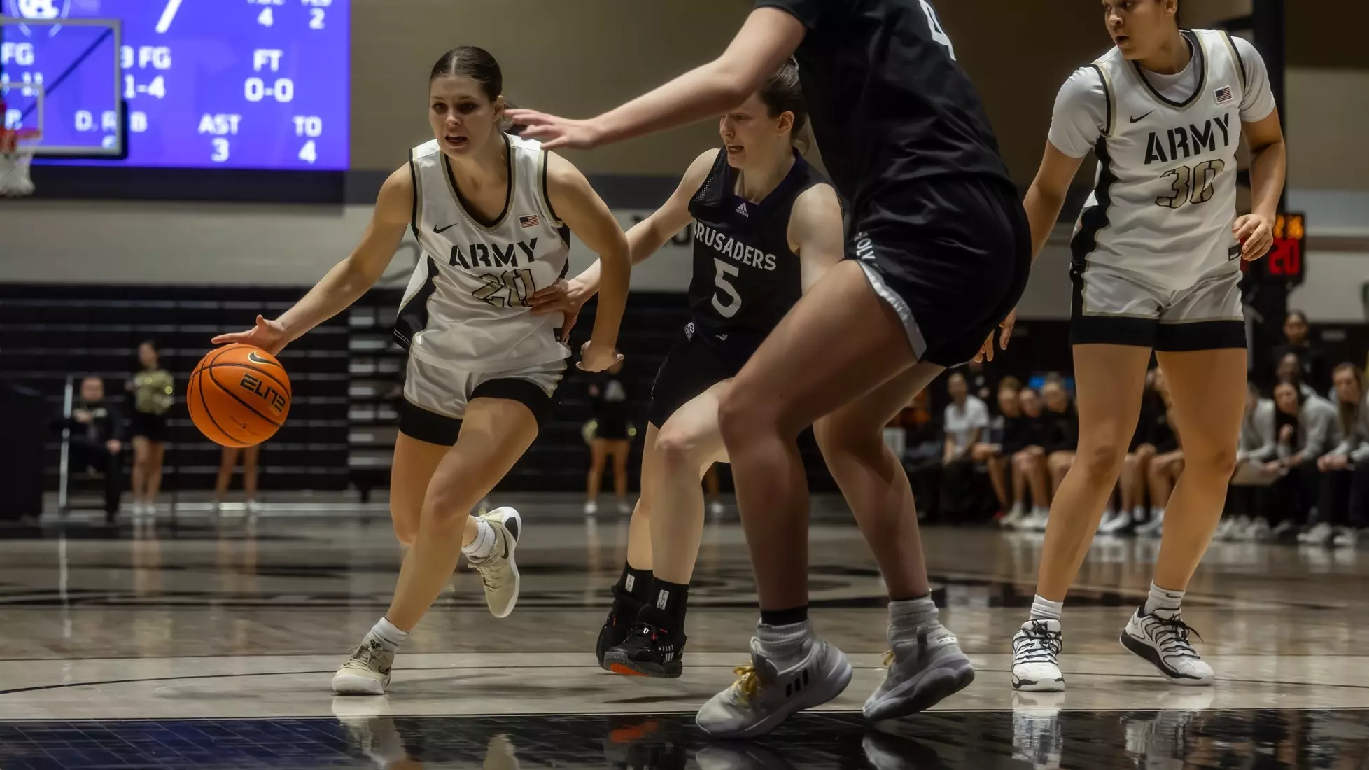 Army Black Knights guard Camryn Tade drives past a defender toward the basket during a home basketball game, wearing a white Army uniform. Teammates and opponents are visible in the background.