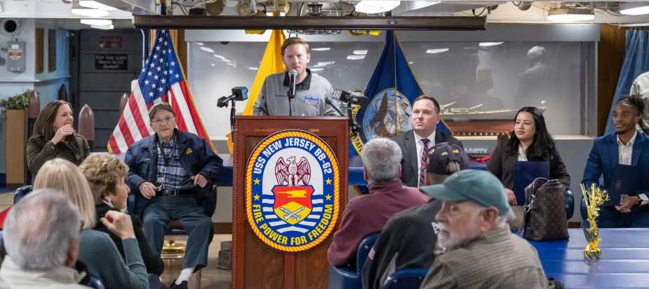 A speaker addresses attendees at a podium during WWII Navy veteran John “Johnny Q” Quinesso Sr.’s 100th birthday recognition ceremony aboard the Battleship New Jersey Museum in Camden, N.J.