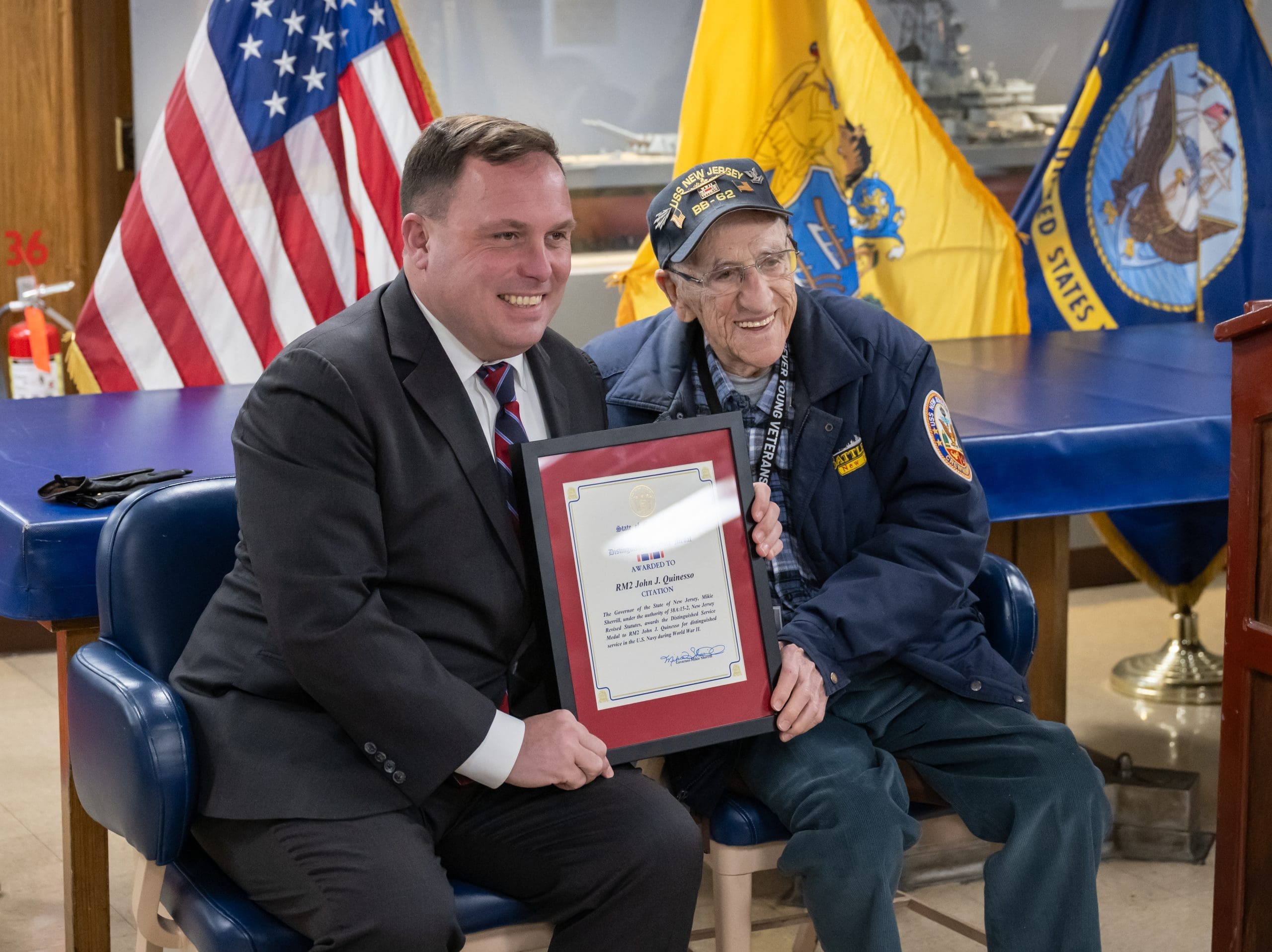 WWII Navy veteran John “Johnny Q” Quinesso Sr. smiles while holding a framed service citation plaque during his 100th birthday celebration aboard the Battleship New Jersey in Camden, N.J.