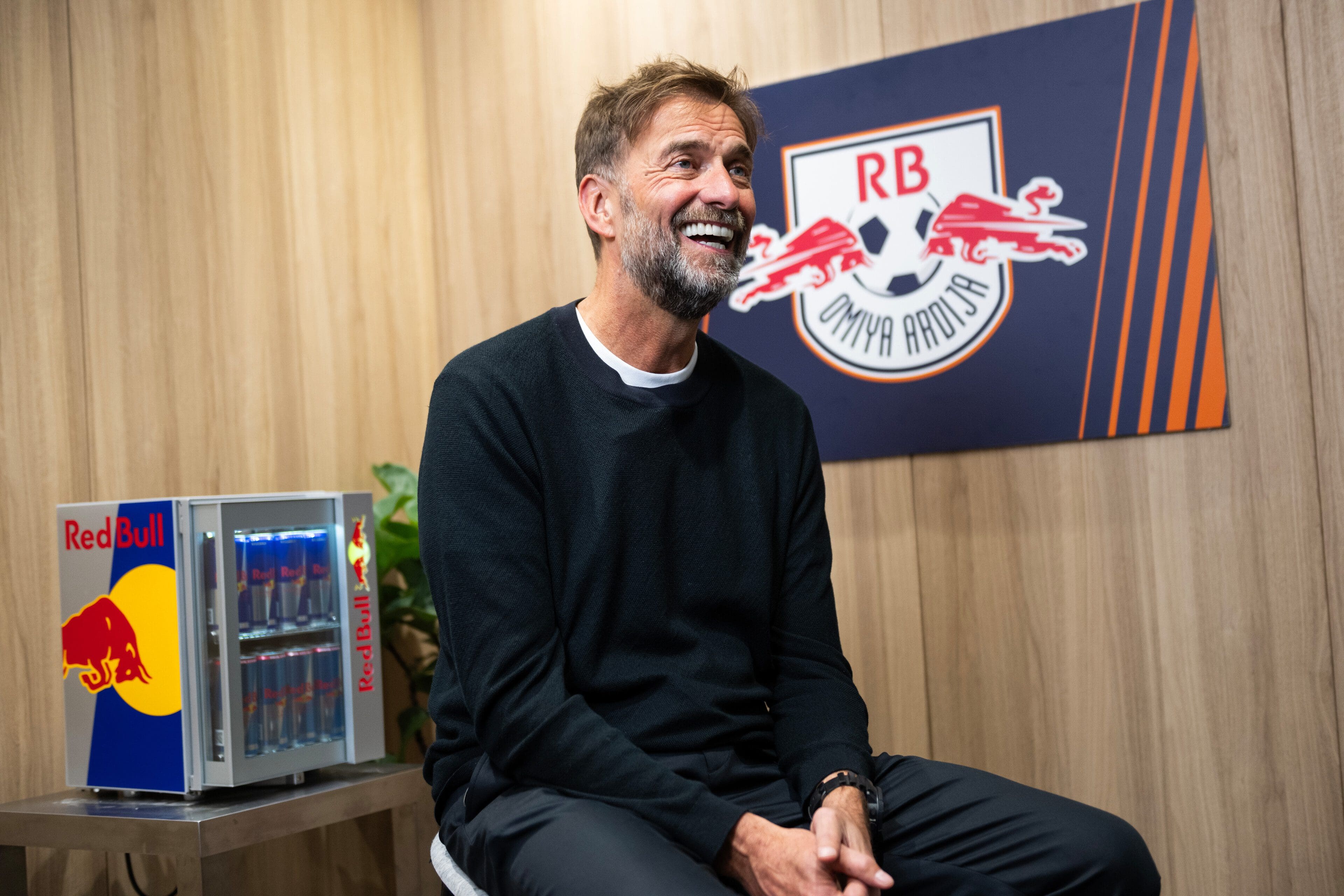 Jurgen Klopp sitting on a stool in an office at RB Salzburg in Austria with the club logo and a red bull mini fridge in the background filled with red bull energy drinks.