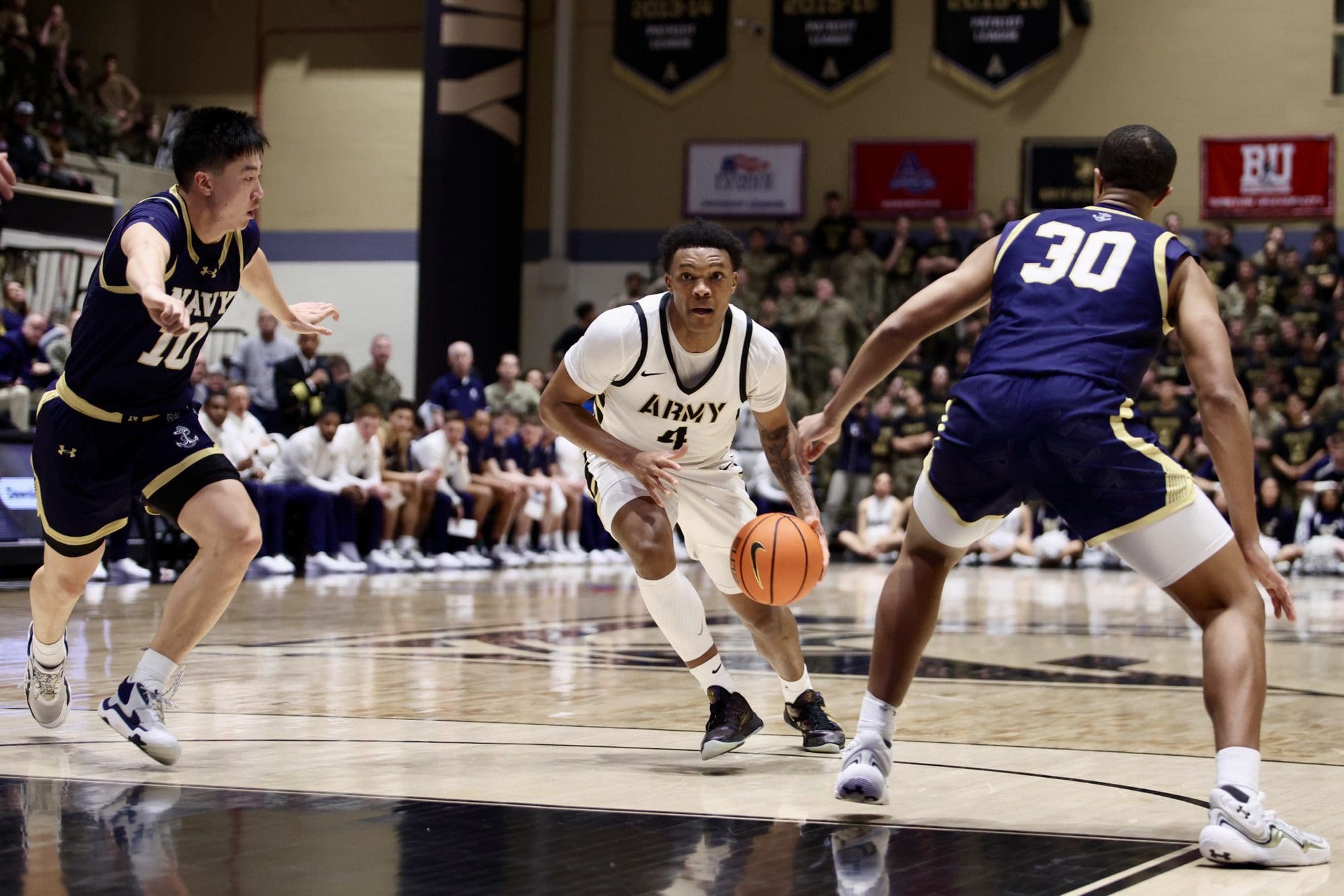 Army guard Jaxson Bell drives upcourt while defended by two Navy players during the Army–Navy rivalry game at Christl Arena.