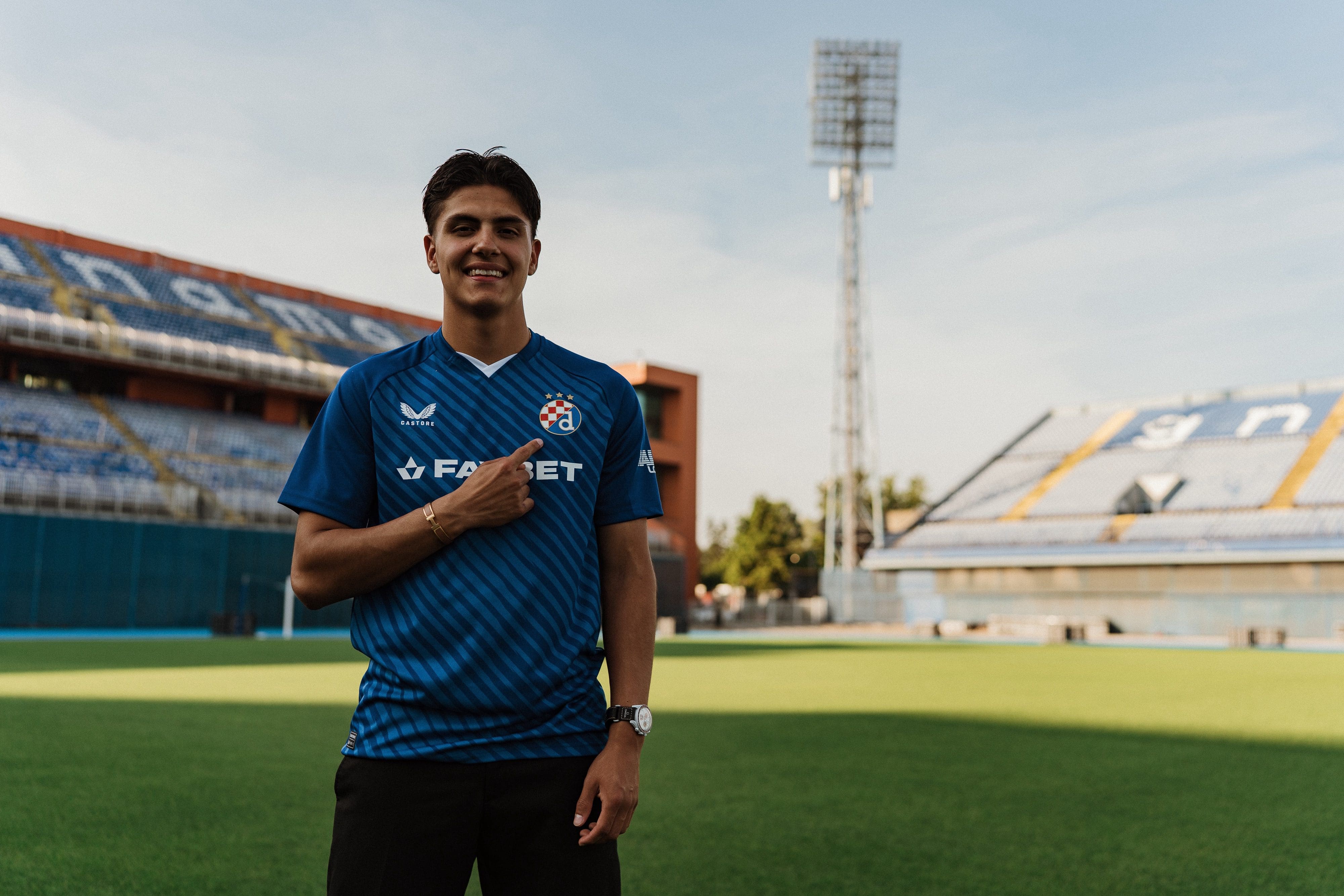 Matteo Pérez-Vinlöf poses on the pitch in a GNK Dinamo Zagreb blue kit, pointing to the club crest at the stadium.