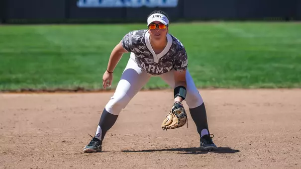 An Army West Point softball player fields in a ready stance on the infield dirt during a game.