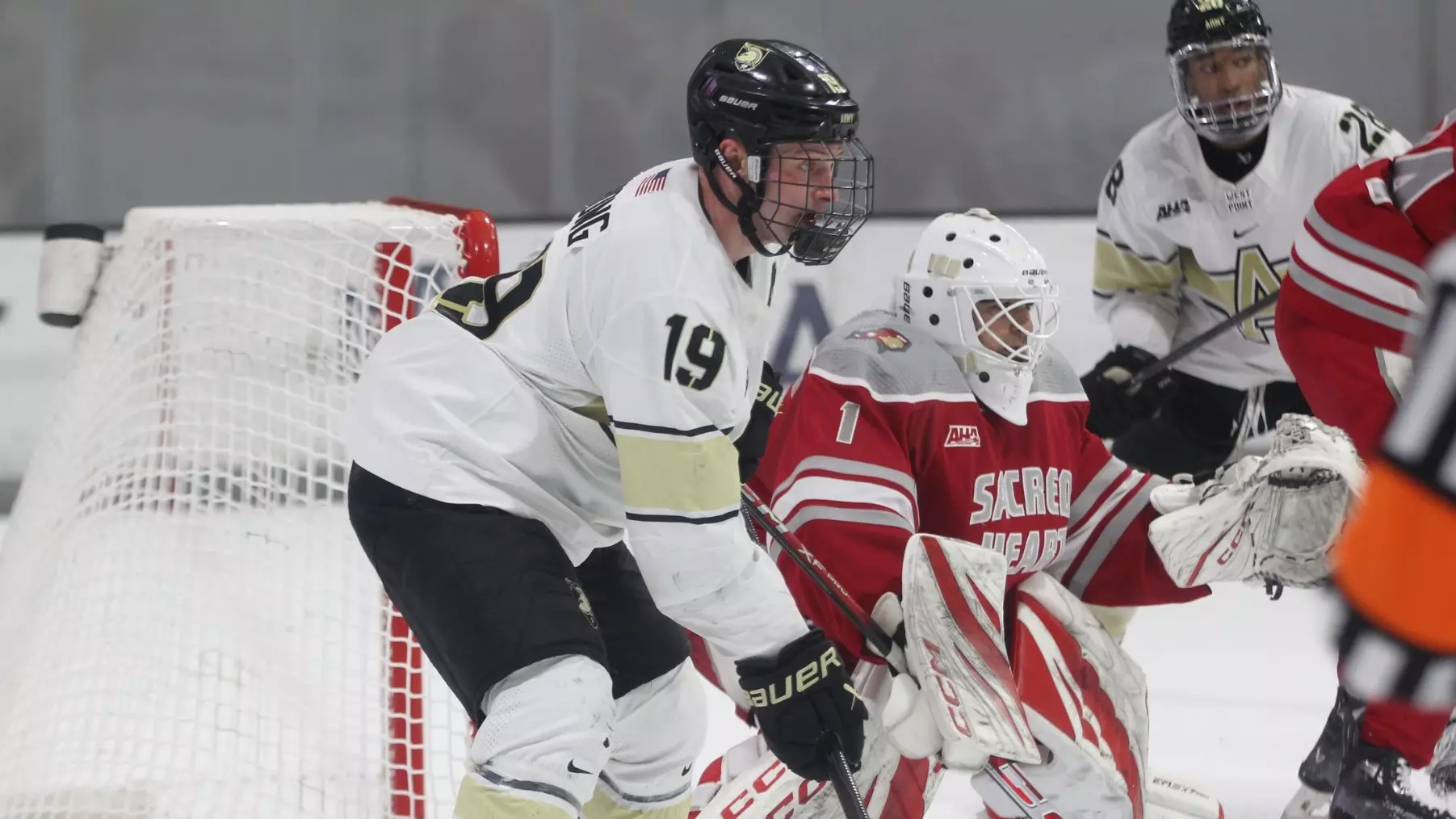 Army West Point hockey player Nik Hong #19 in a white jersey battling for position in front of the net while a Sacred Heart goaltender defends.