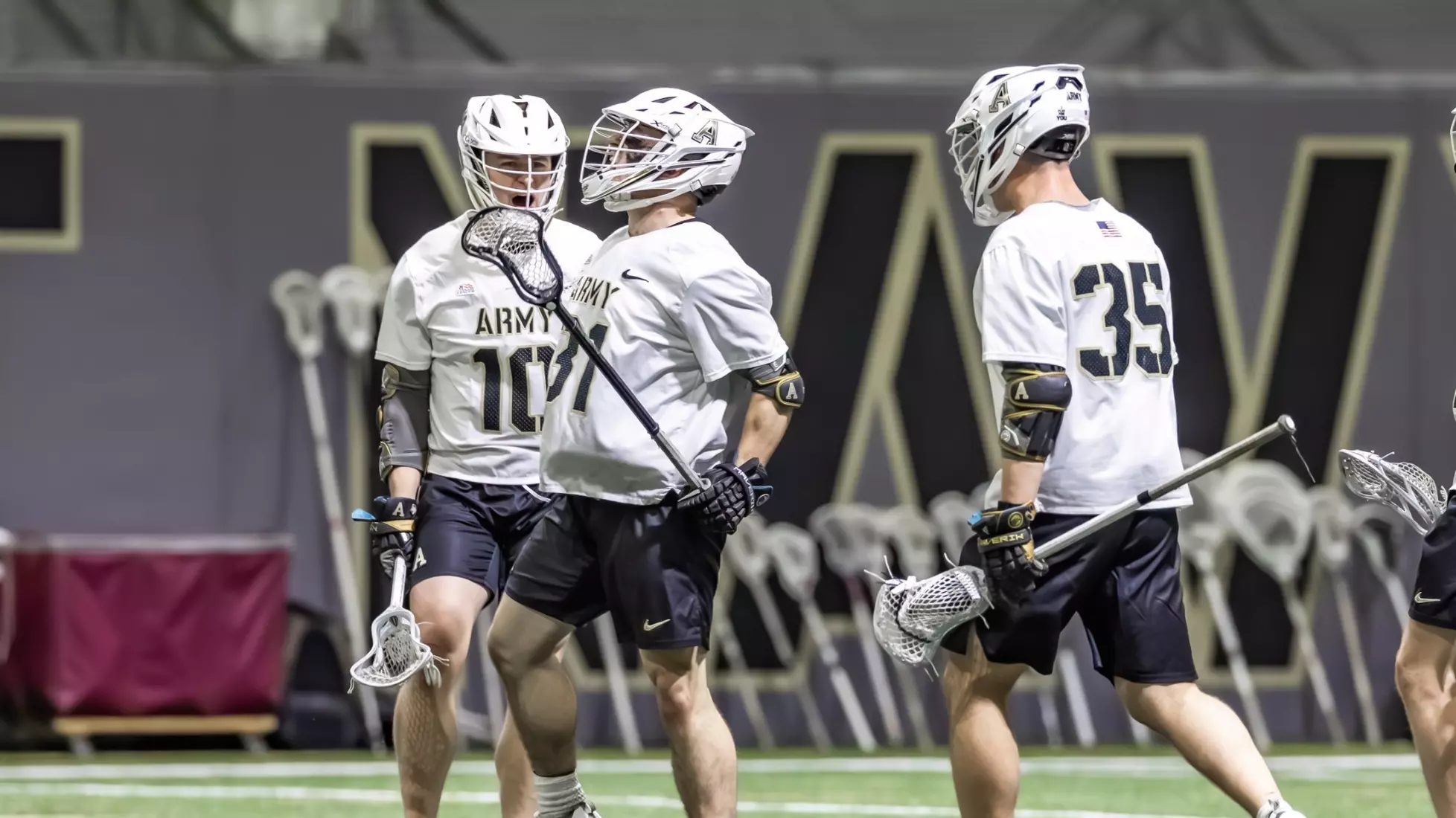 Three Army West Point men’s lacrosse players in white practice jerseys and helmets stand on the field during a training session.