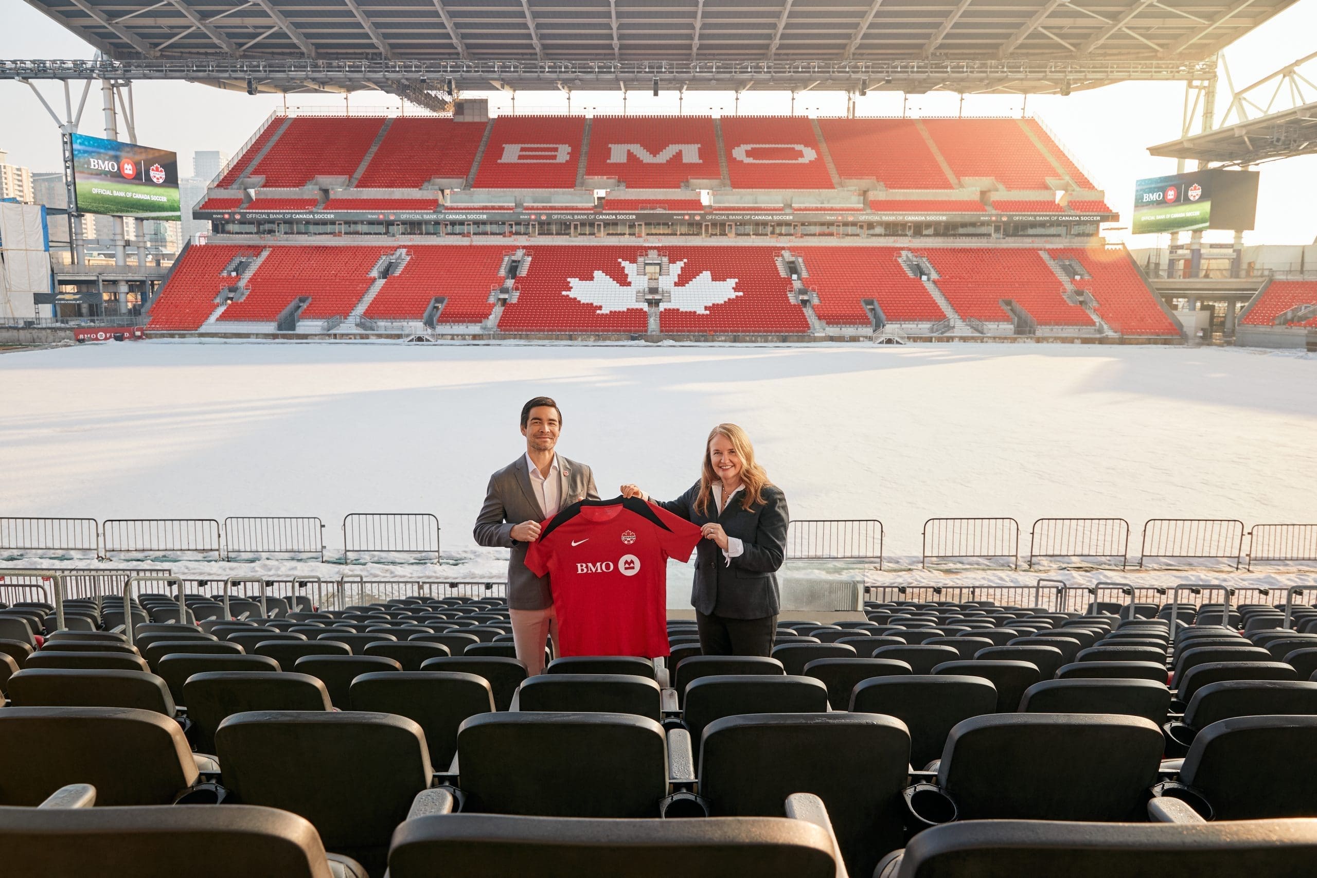 Canada Soccer CEO Kevin Blue and BMO CMO Catherine Roche standing in the empty stands of BMO Field, holding up a red Nike training jersey featuring the BMO logo on the chest.