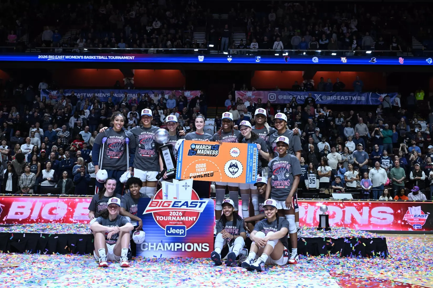UConn players celebrate on a confetti‑covered court after winning the 2026 Big East Women’s Basketball Tournament, holding the trophy and a “2026 Ticket Punched” sign.