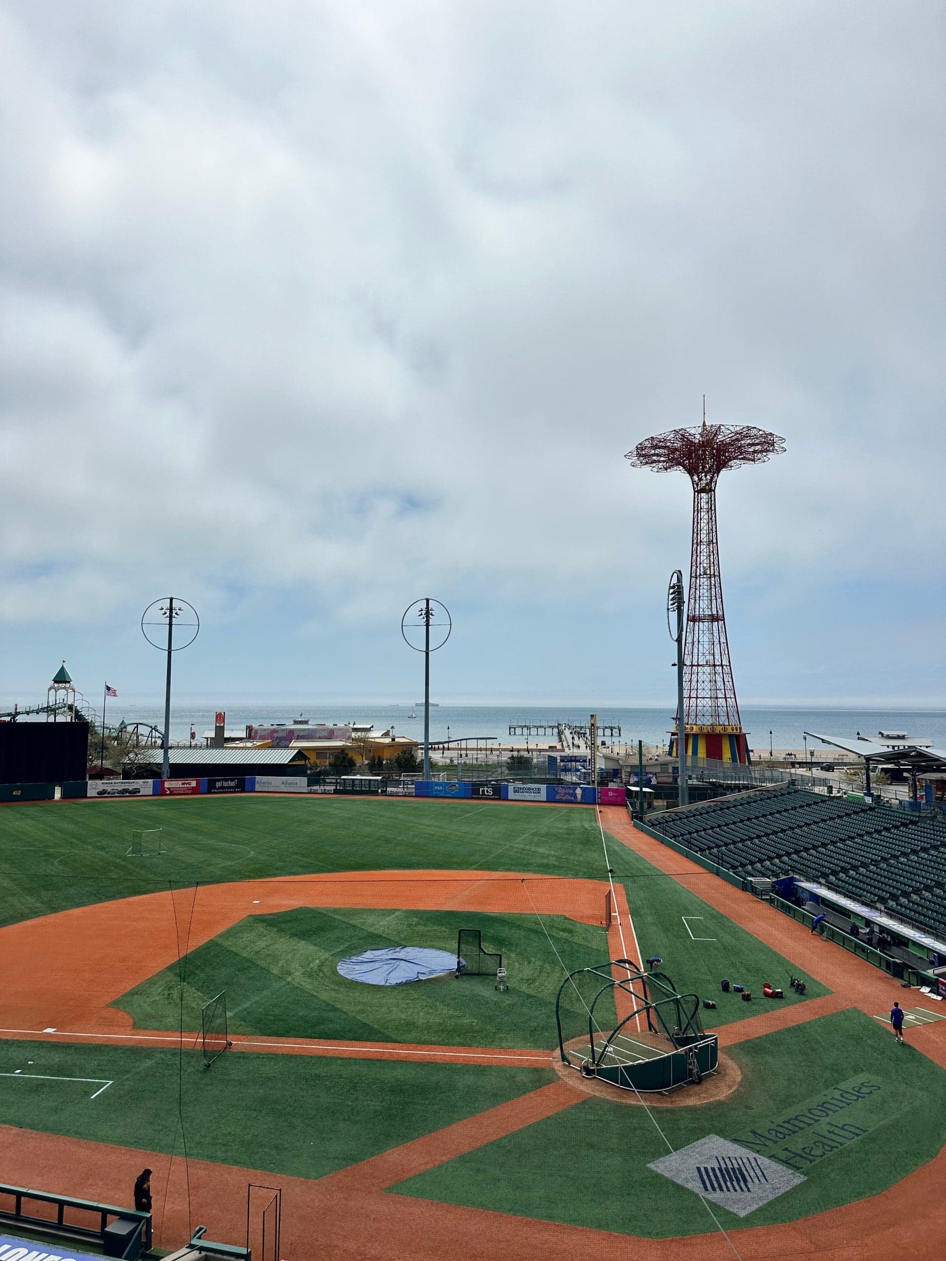A wide view of Maimonides Park during pregame batting practice, with the Coney Island boardwalk, Parachute Jump, and ocean visible beyond the outfield under a cloudy sky.