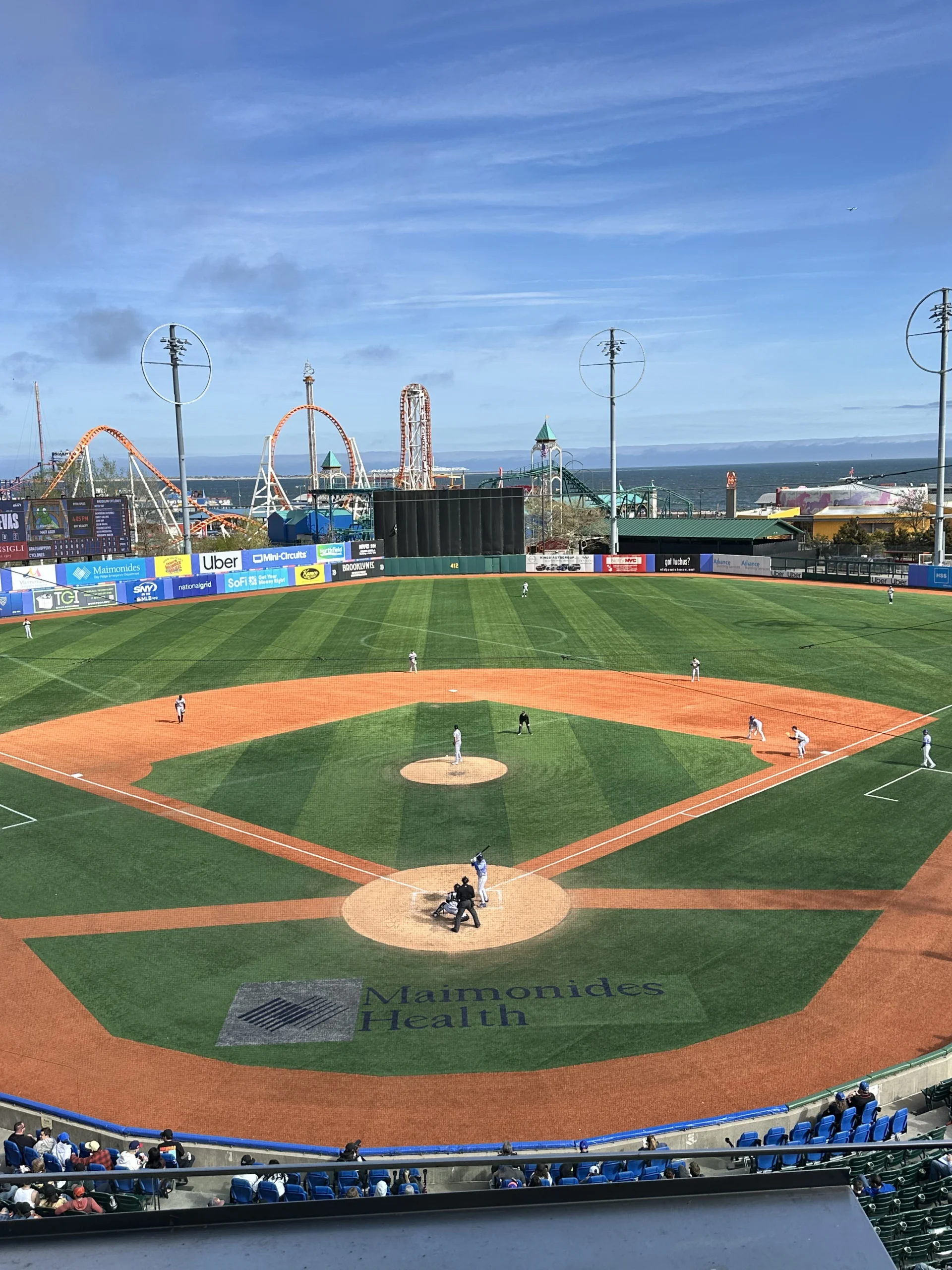 Daytime baseball game at Maimonides Park viewed from behind home plate, with the batter set in the box and Coney Island’s amusement rides and the ocean visible beyond the outfield.