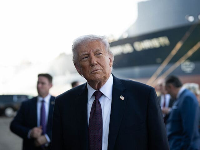 President Donald J. Trump stands outdoors in a dark suit, white shirt, and red tie during an official appearance at a port facility, with staff and security personnel visible in the background.